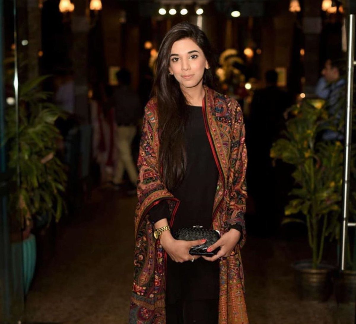 A woman wearing a black dress and a patterned shawl posing outdoors at night.