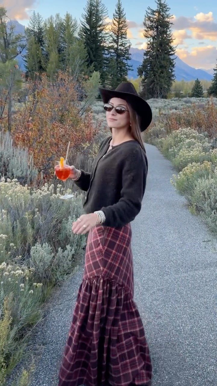 Woman in a black cowboy hat, black cardigan, and burgundy plaid skirt holding a cocktail outdoors.