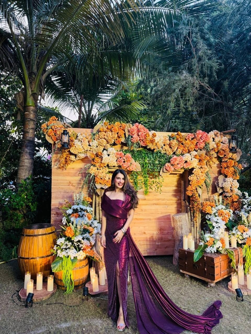 A woman wearing a long, sleeveless, shimmering purple gown standing in a floral garden setting.