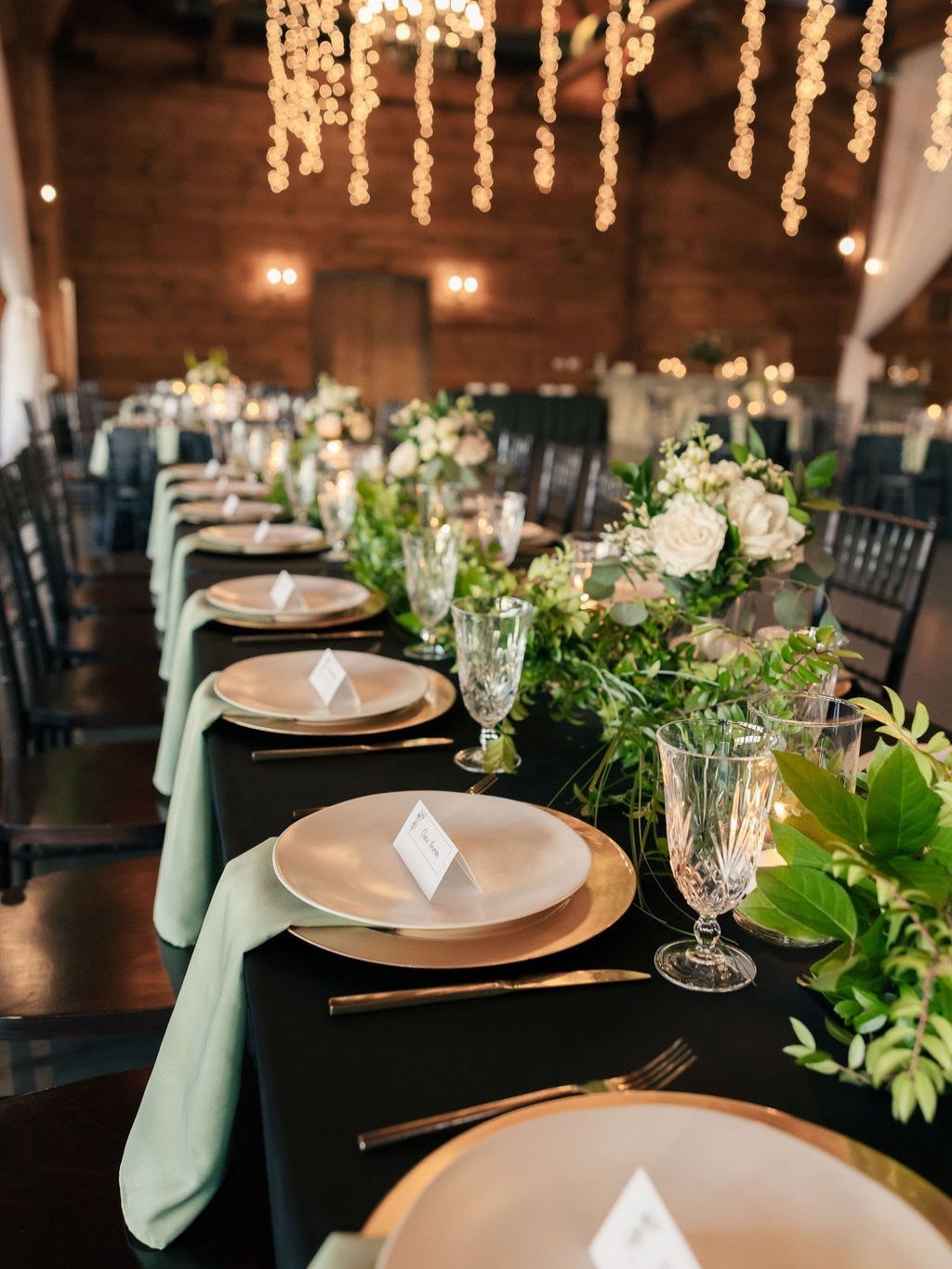 A long wedding reception table decorated with white flowers, green foliage, and gold-rimmed plates under hanging lights.