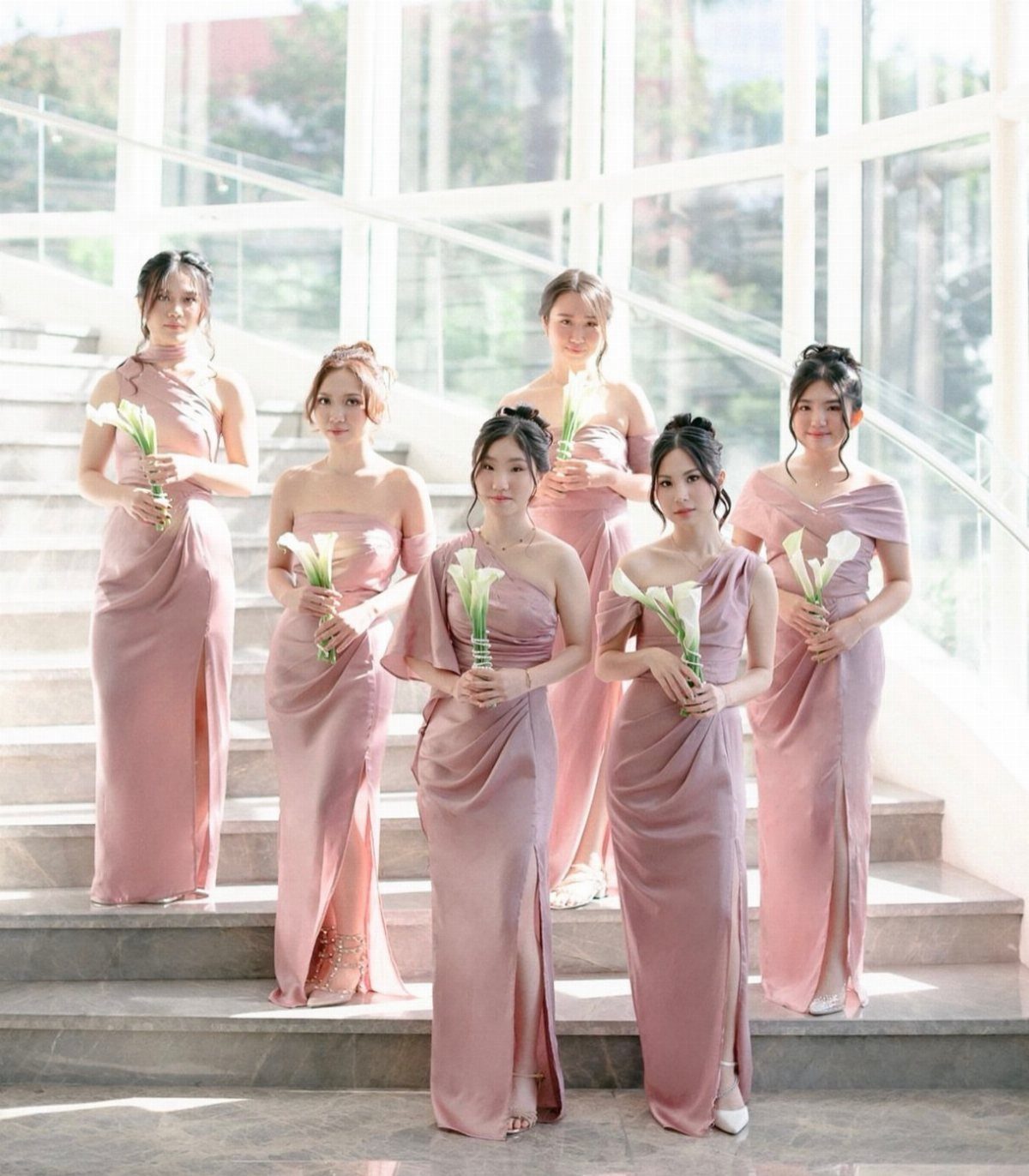 Six bridesmaids in blush pink satin dresses holding white calla lily bouquets on stone stairs.