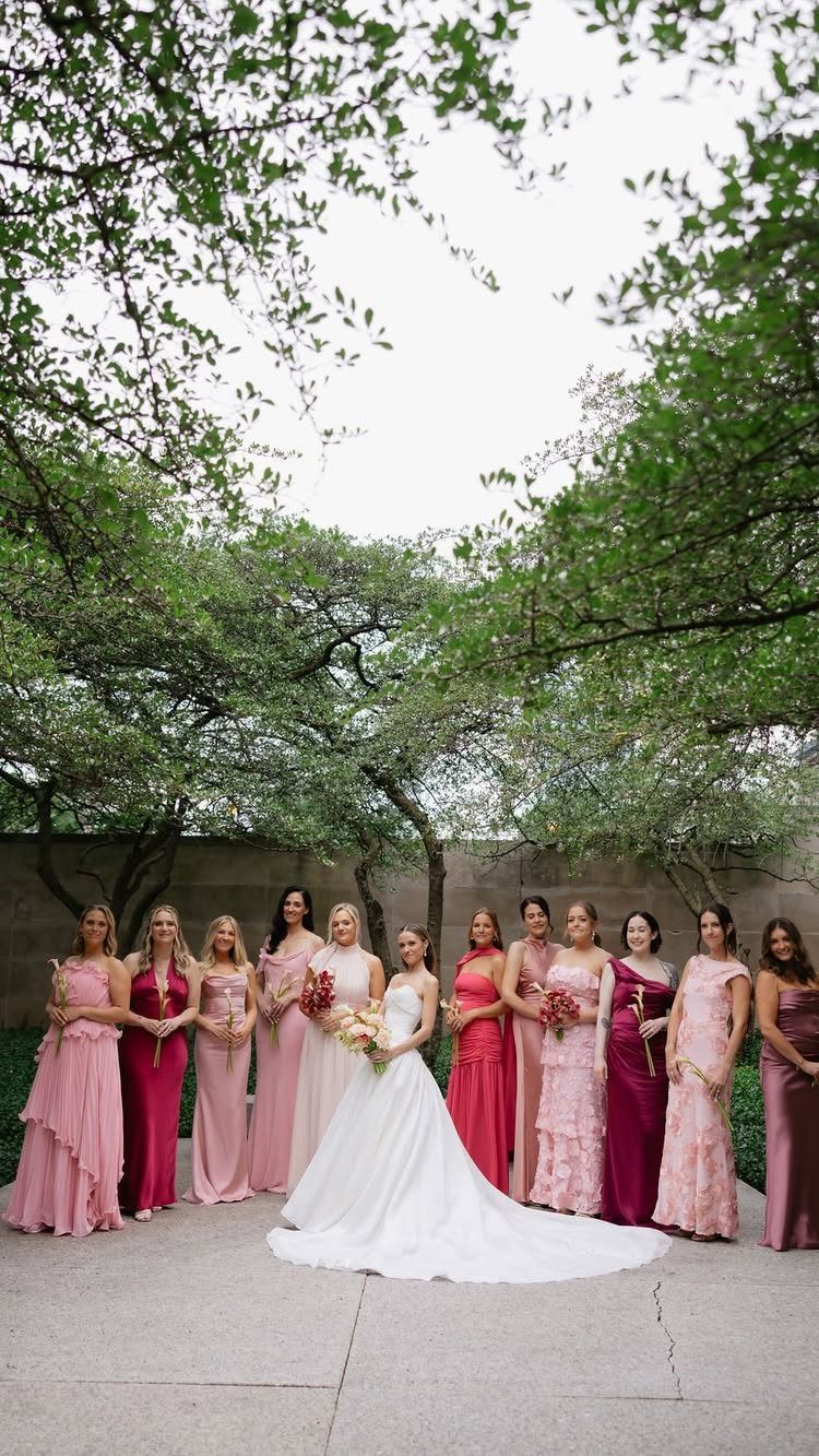 A bride in white stands with bridesmaids wearing pink and burgundy dresses in a garden.