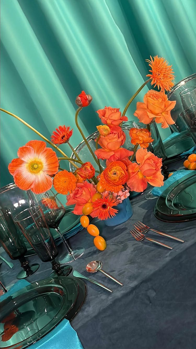 Orange flowers in a blue vase set on a dark blue table with teal glassware.