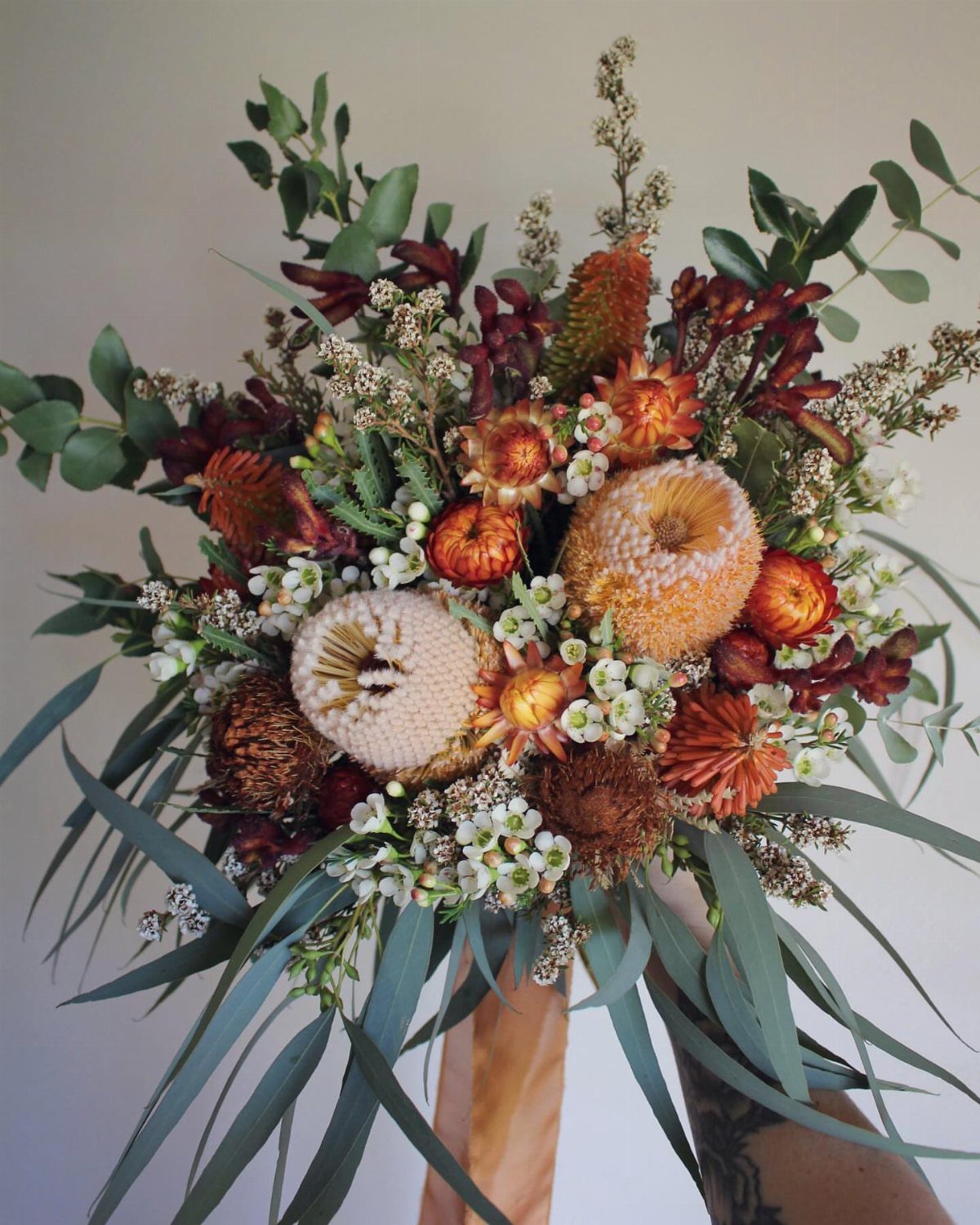 A large bouquet of orange and brown proteas and eucalyptus held by a tattooed hand.