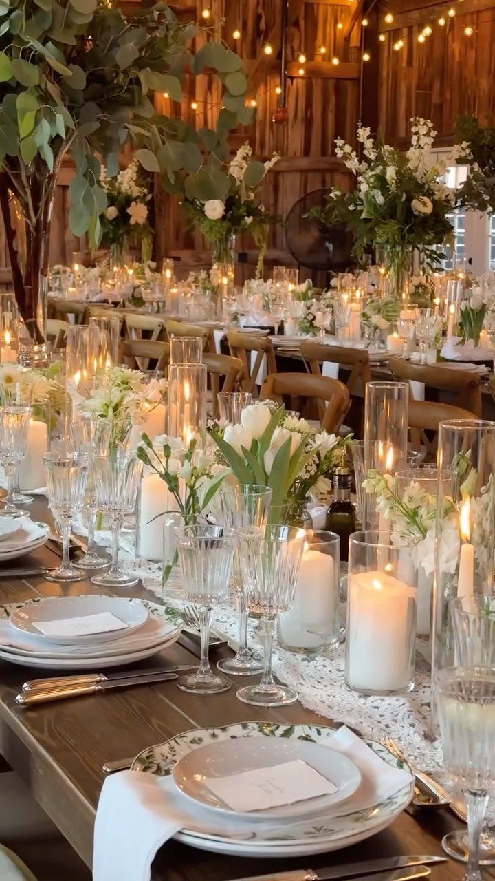A long rustic wedding table decorated with white flowers, tall candles, and elegant glassware in a barn.