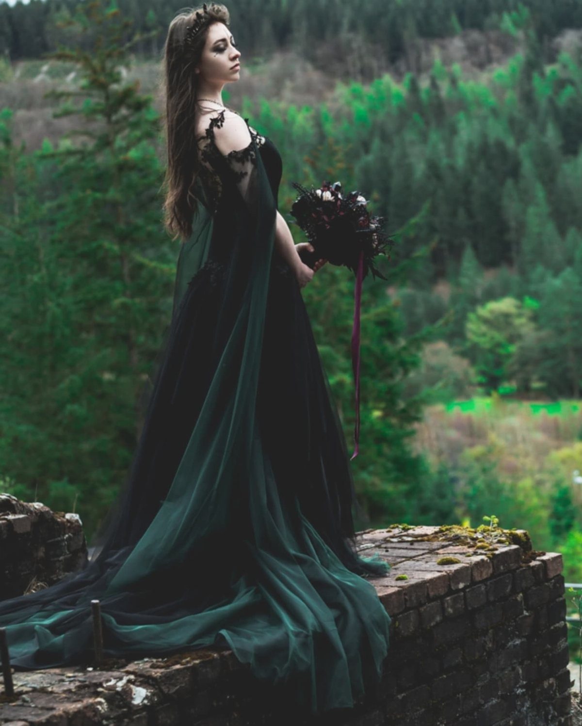 7 Enchanting Ideas for Witchy Wedding Dresses 2 A woman in a dark green and black tulle gown holding a bouquet in a forest.